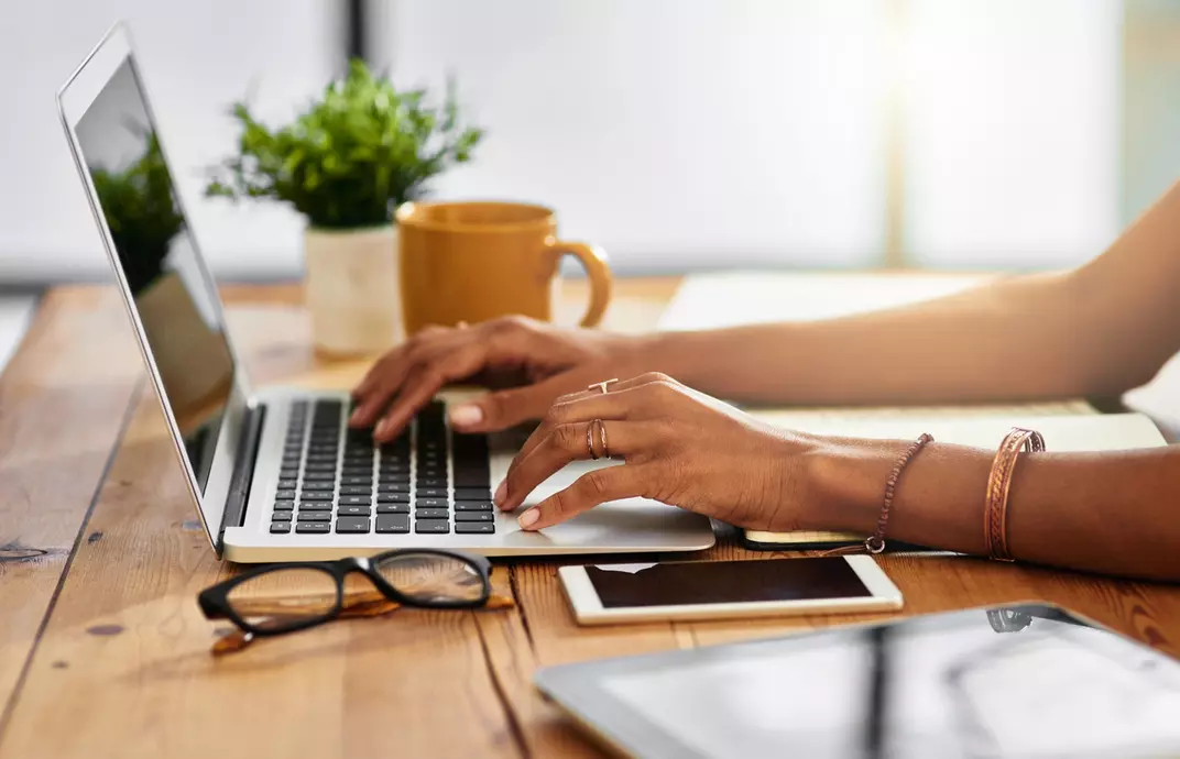 Mão de uma mulher digitando algo no laptop que está apoiado sobre a mesa com mais alguns objetos