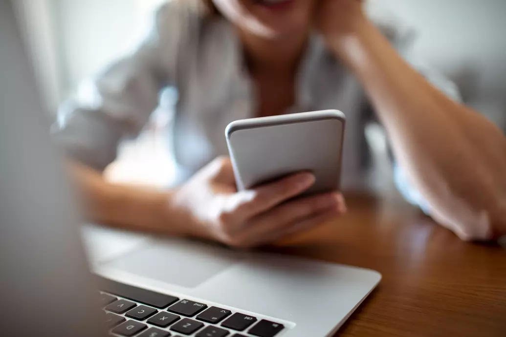 Mulher sentada a mesa segurando e olhando para o celular, próxima ao laptop