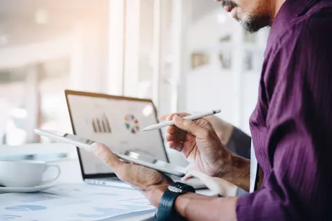Homem vestindo camisa social roxa usando tablet e computador com gráfico financeiro