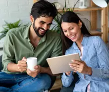 Homem segura uma caneca e sorri ao lado de uma mulher em uma sala de estar. Ela também sorri enquanto segura um tablet. Ambos estão olhando para ele.